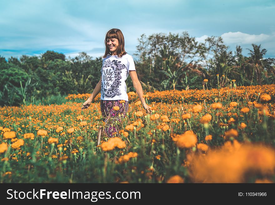 Woman in White and Black Floral Crew-neck T-shirt and Red Bottoms Standing on Orange Petaled Flower Field at Daytime