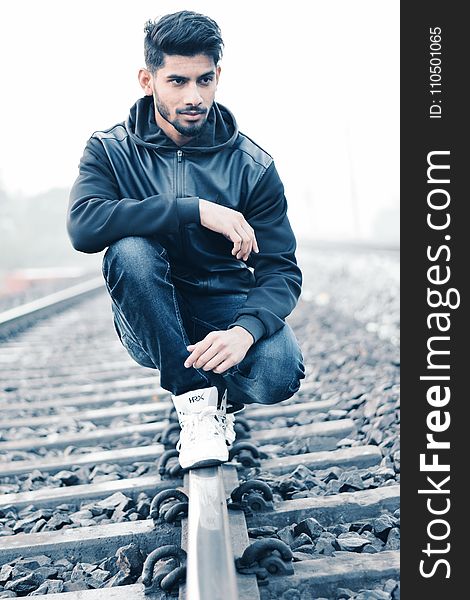 Man in Black Leather Zip-up Hoodie and Black Denim Jeans Sitting on Grey Metal Train Railings Surrounded With Rocks during Husky M