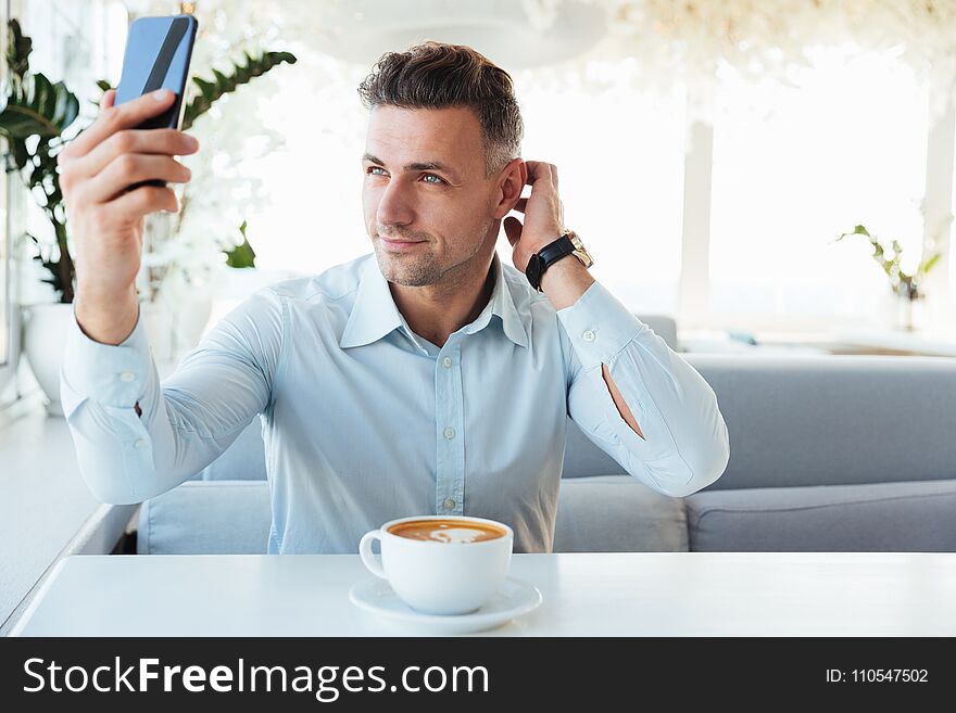 Smiling caucasian mature man resting alone in cafe with cup of cappuccino and making selfie on black smartphone