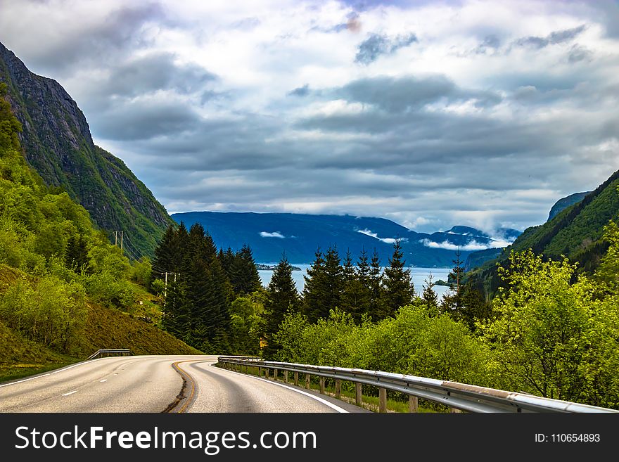 Grey Pathway Surrounded By Green Trees On Both Sided Leading To Blue Moutains Under Grey Cloudy Sky