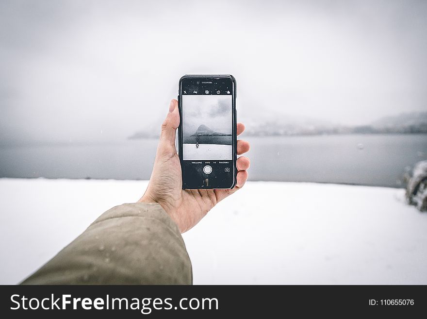 Photo Of Person Wearing Brown Coat Holding Android Smartphone While Taking Picture Of Mountain And Body Of Water