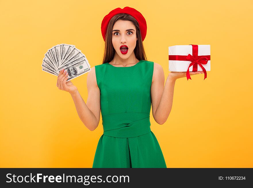 Portrait of a shocked young woman dressed in beret and green dress showing bunch of money banknotes and holding gift box over yellow background