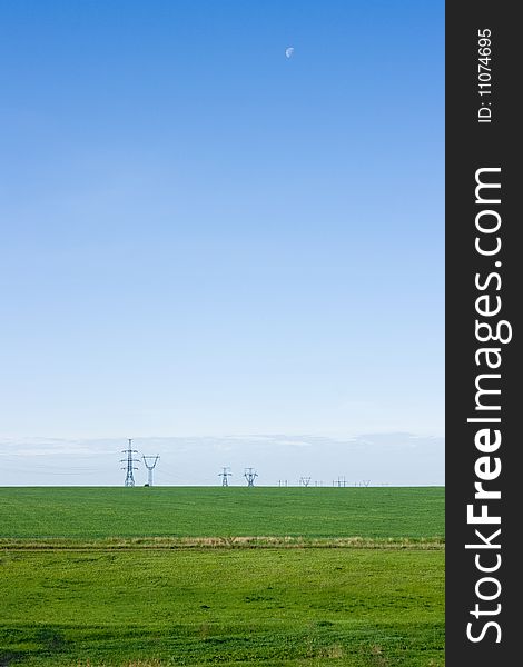 Green field and blue sky. Moon in the sky. Power lines are visible on the horizon.