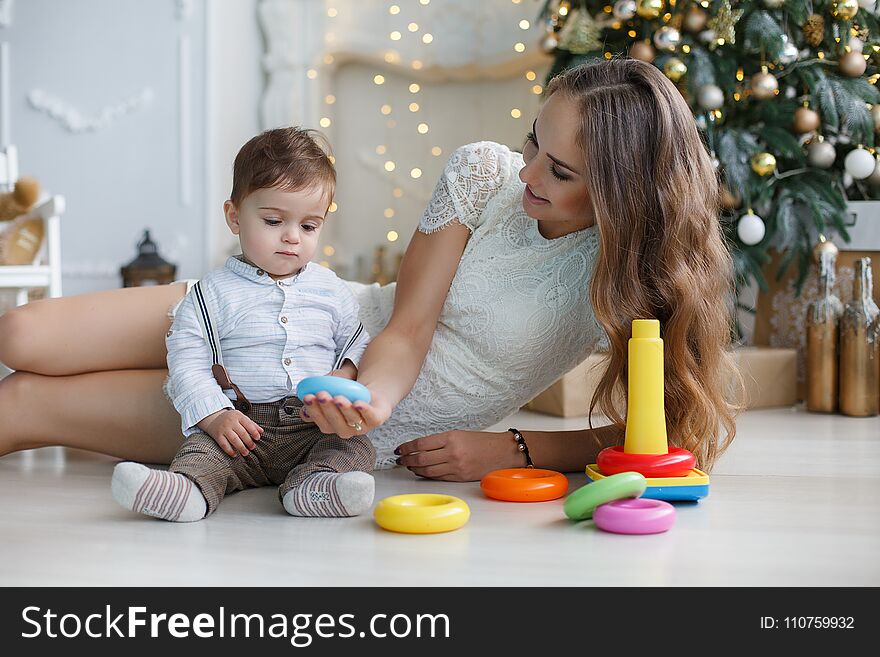 Young women with long hair,dressed in a short white lace dress lies on white floor with sitting next to a young son,dressed in a white shirt and grey trousers,mother teaches son how to assemble a pyramid,the back is green decorated Christmas tree