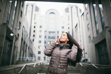 Young Girl Talking On Mobile Phone In Courtyard Business Center. Girl With Long Dark Hair Dressed In Winter Jacket In Cold Weather Royalty Free Stock Images Young Girl Talking On Mobile Phone In Courtyard Business Center. Girl With Long Dark Hair Dressed In Winter Jacket In Cold Weather Royalty Free Stock Images