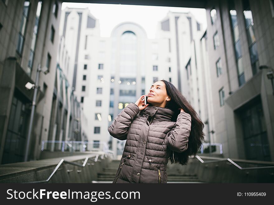 Young girl talking on mobile phone in courtyard business center. girl with long dark hair dressed in winter jacket in cold weather speaks on phone on background buildings made of glass and concrete.