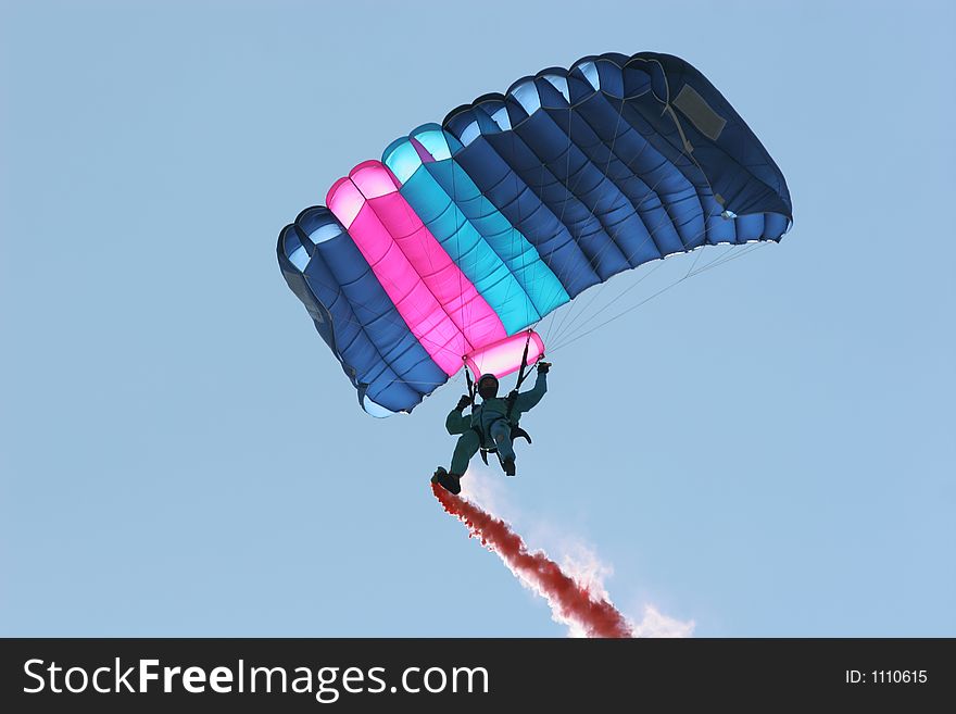 Parachutist in a blue boiler suit, with a pink and blue parachute, flying through the air with a red smoke trail coming from his right foot. Parachutist in a blue boiler suit, with a pink and blue parachute, flying through the air with a red smoke trail coming from his right foot..
