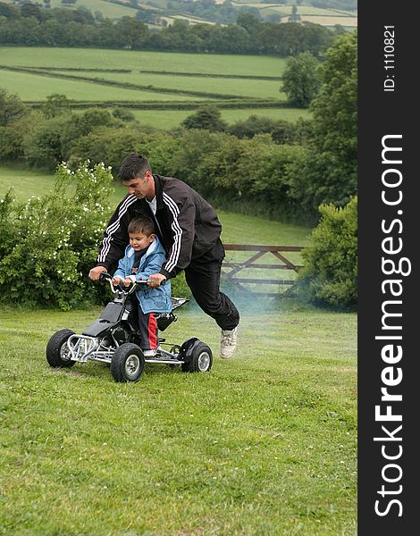 Little boy on racing a mini moto quad bike with his father riding on the back. Set in countryside. Little boy on racing a mini moto quad bike with his father riding on the back. Set in countryside.