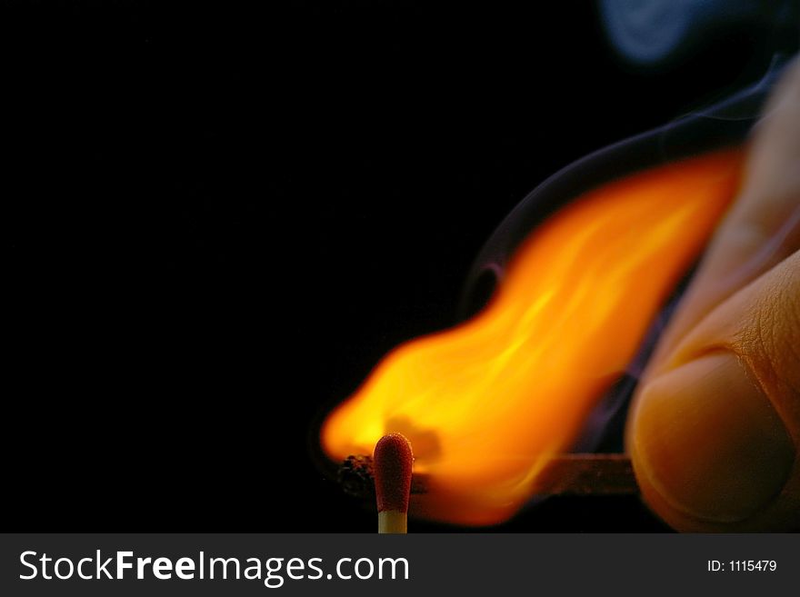 Closeup of a red-tipped wooden match stick at ignition from another match being held by fingers. Red, orange, yellow, and white flame. Black background. Closeup of a red-tipped wooden match stick at ignition from another match being held by fingers. Red, orange, yellow, and white flame. Black background.