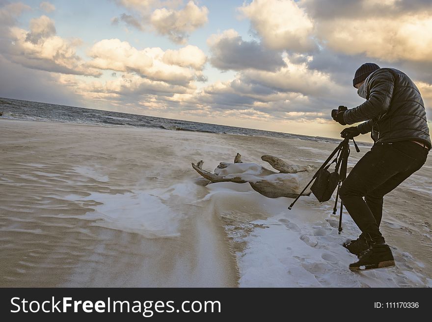 Man in Black Bubble Jacket Walking on Seashore While Holding Black Camera Tripod