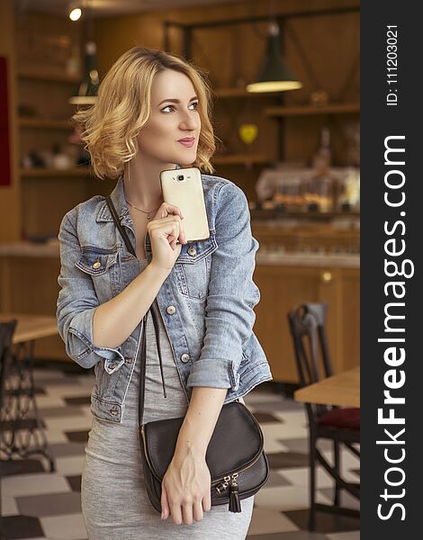 Blonde with dark roots caucasian woman in casual summer outfit at the cafe. Grey dress and jeans jacket. Woman got natural day makeup and curly hairstyle. She talks on her phone, working, make calls