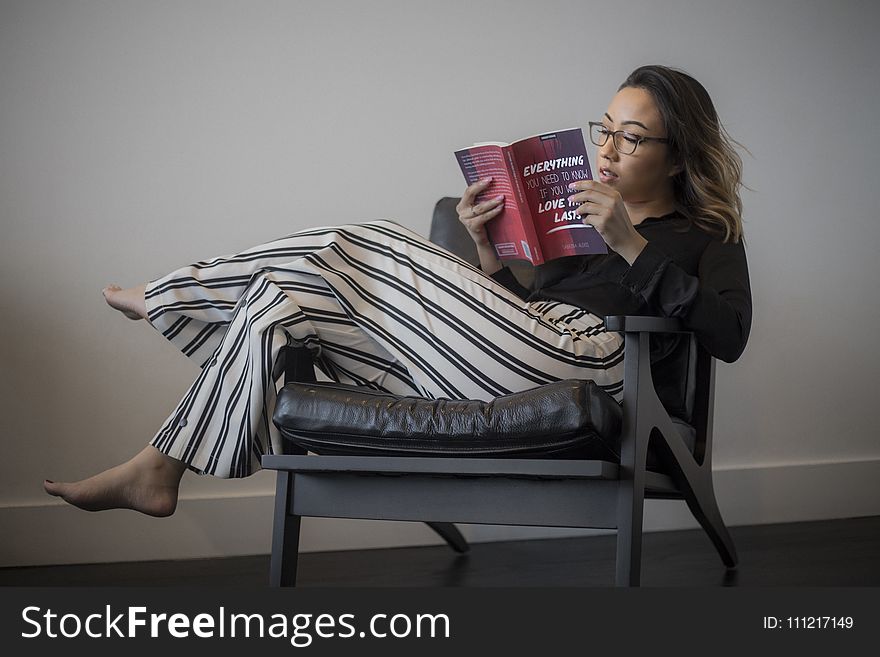 Women&x27;s Black Long-sleeved Top, White And Black Striped Pants Reading Book Sitting On Gray Wooden Framed Padded Armchair