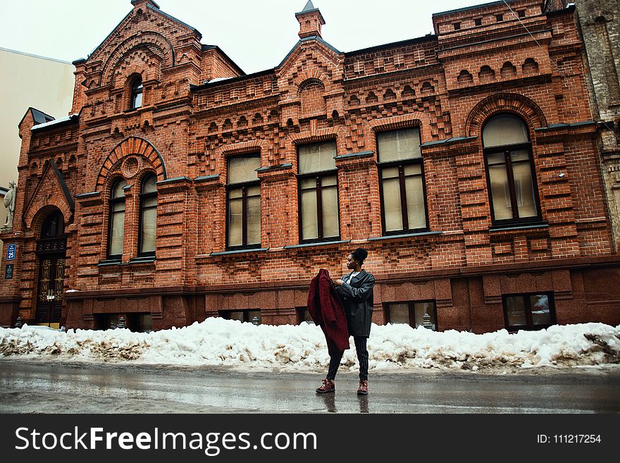 Man Holding Red Coat Standing on Street Near Brown Building