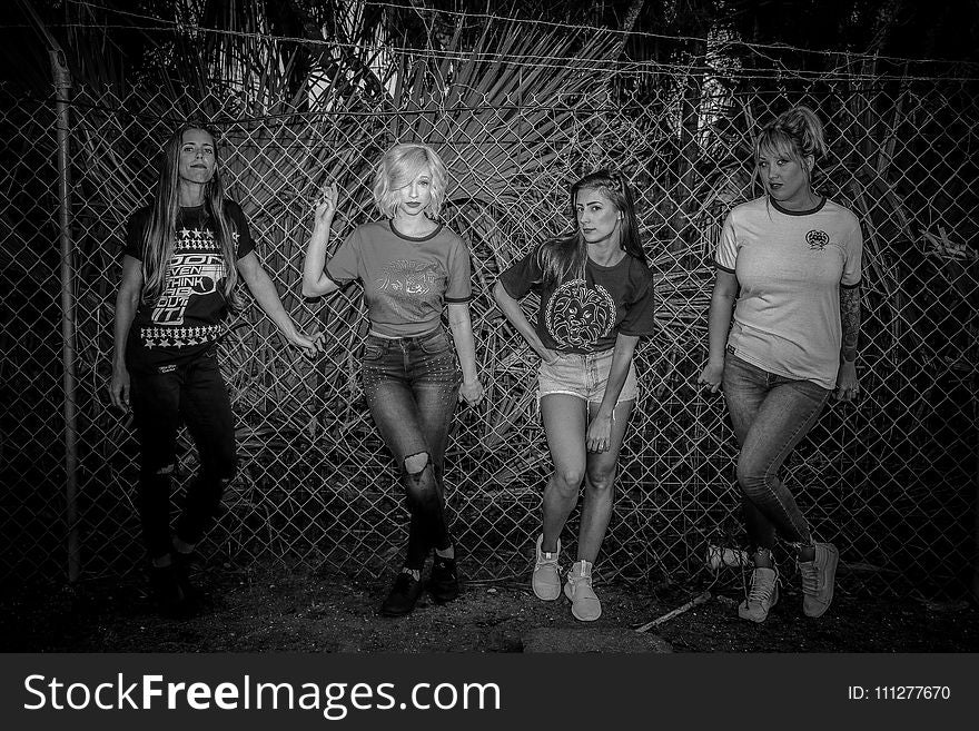Grayscale Photo of Four Woman Standing Near on Link Fence