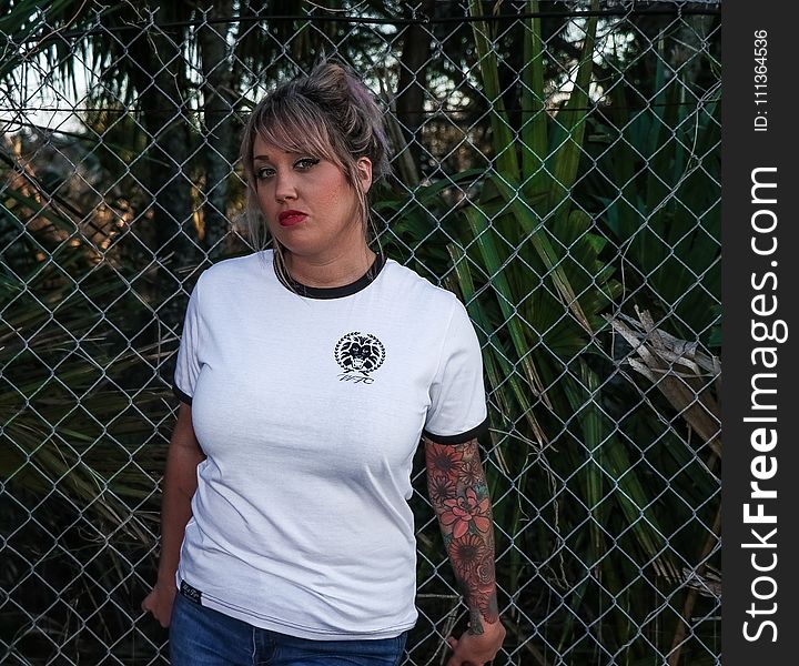 Female Wearing Black And White Crew-neck T-shirt And Blue Denim Bottoms Leaning On Chicken Wire