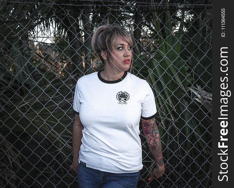 Woman Wearing White Crew-neck T-shirt With Black Trim and Blue Denim Bottoms Beside Wire Fence