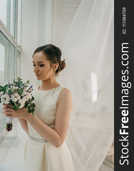 Woman in White Sleeveless Gown Holding White Flower Bouquet Infront of Window