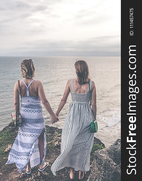 Two Women Wearing Sleeveless Dress Holding Each Others Hand On Cliff