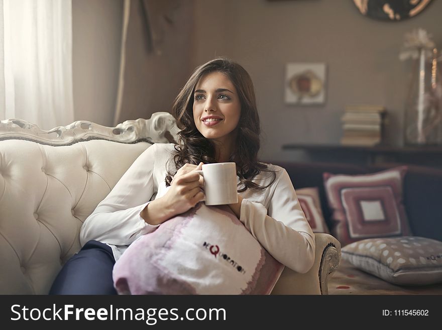 Woman Wearing White Top Drinking Beverage From White Ceramic Mug While Lying On Sofa Inside Well Lit Room