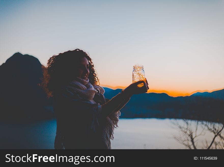Woman Holding Mason Jar With String Light With Lake and Mountain over View during Golden Hour