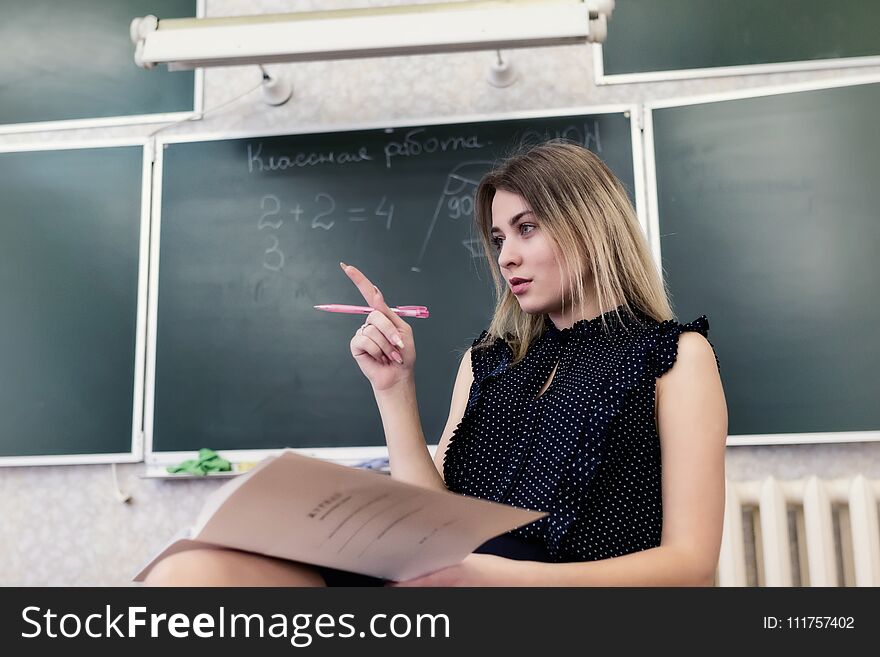 Strict young blonde teacher sits on a chair in the classroom against a blackboard and reflects on who to check homework. Strict young blonde teacher sits on a chair in the classroom against a blackboard and reflects on who to check homework