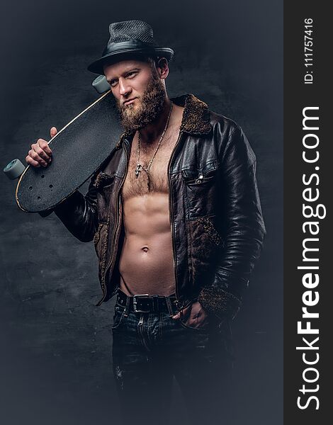 Bearded male dressed in leather jackett holds skateboard.