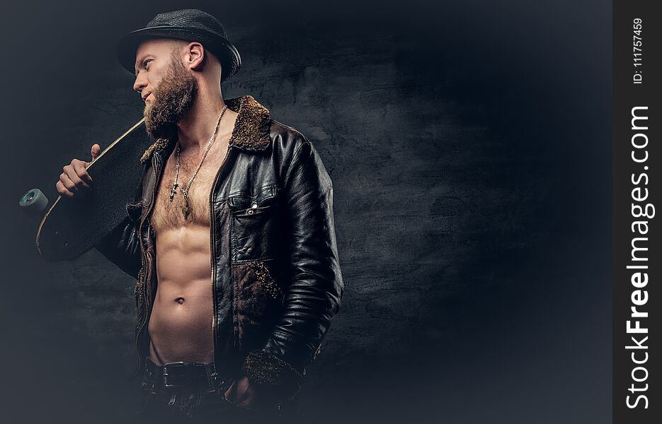 Bearded male dressed in leather jackett holds skateboard.