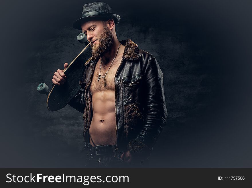 Bearded male dressed in leather jackett holds skateboard.