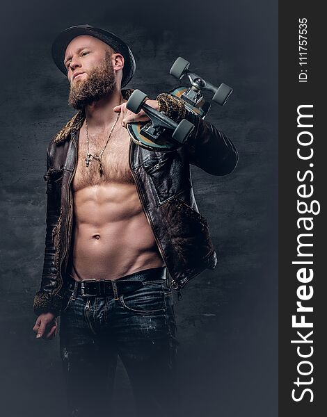 Bearded male dressed in leather jackett holds skateboard.