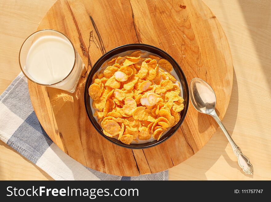 Sweet cornflakes breakfast cereal in the bowl with glass of milk on the wooden table and spoon. Scene lit with direct sun rays. Horizontal image. Selective focus. Sweet cornflakes breakfast cereal in the bowl with glass of milk on the wooden table and spoon. Scene lit with direct sun rays. Horizontal image. Selective focus