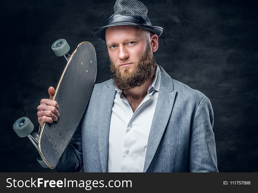 Stilysh bearded male in a suit and felt hat holds skateboard on a shoulder. Stilysh bearded male in a suit and felt hat holds skateboard on a shoulder.