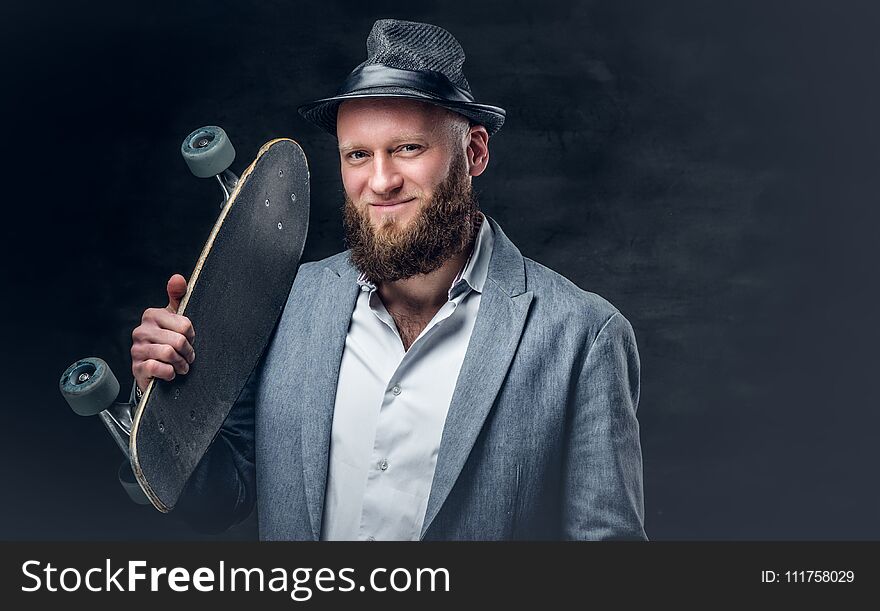 Stilysh bearded male in a suit and felt hat holds skateboard on a shoulder. Stilysh bearded male in a suit and felt hat holds skateboard on a shoulder.