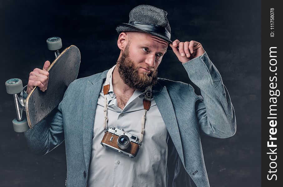 Stylish bearded male in a suit and felt hat holds skateboard and the vintage SLR photo camera. Stylish bearded male in a suit and felt hat holds skateboard and the vintage SLR photo camera.