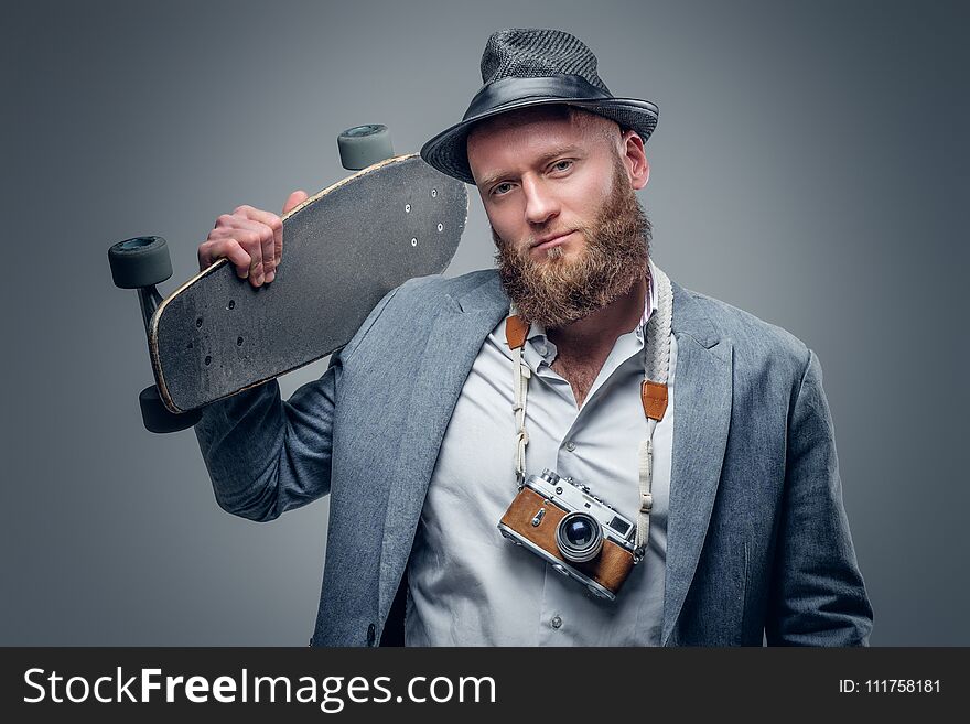 Portrait of a stylish bearded male in a suit and felt hat holds skateboard and vintage SLR photo camera in the grey vignette background. Portrait of a stylish bearded male in a suit and felt hat holds skateboard and vintage SLR photo camera in the grey vignette background.