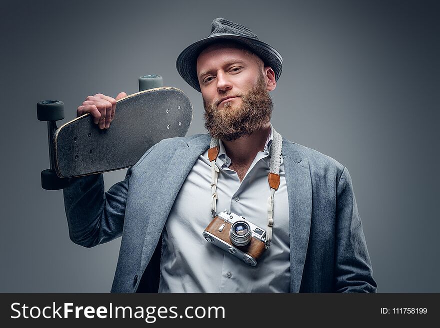 Bearded man in a suit holds skateboard and SLR photo camera.