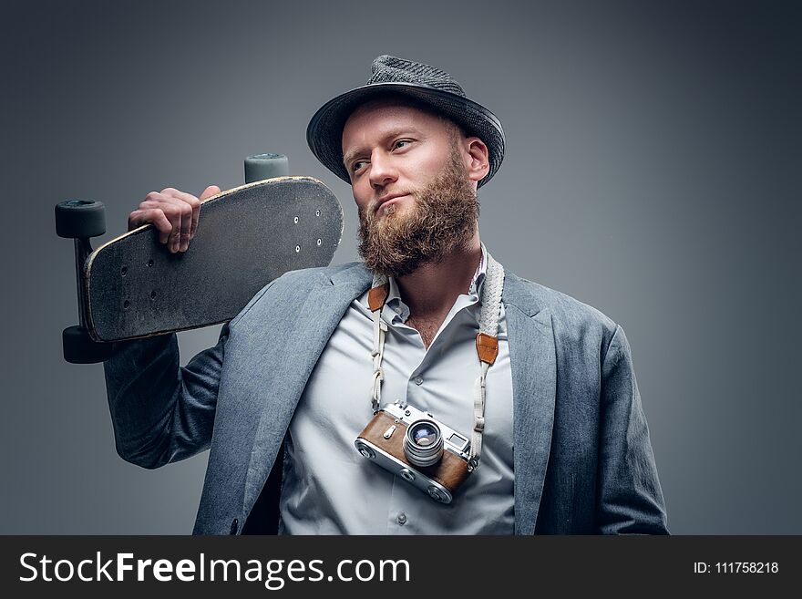 Portrait of a stylish bearded male in a suit and felt hat holds skateboard and vintage SLR photo camera in the grey vignette background. Portrait of a stylish bearded male in a suit and felt hat holds skateboard and vintage SLR photo camera in the grey vignette background.