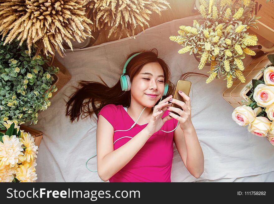 Happy Young Woman Laying on the Floor to Listening Music via Smart Phone. Girl relaxing in Cozy Place, Surrounded by Flower, Top View