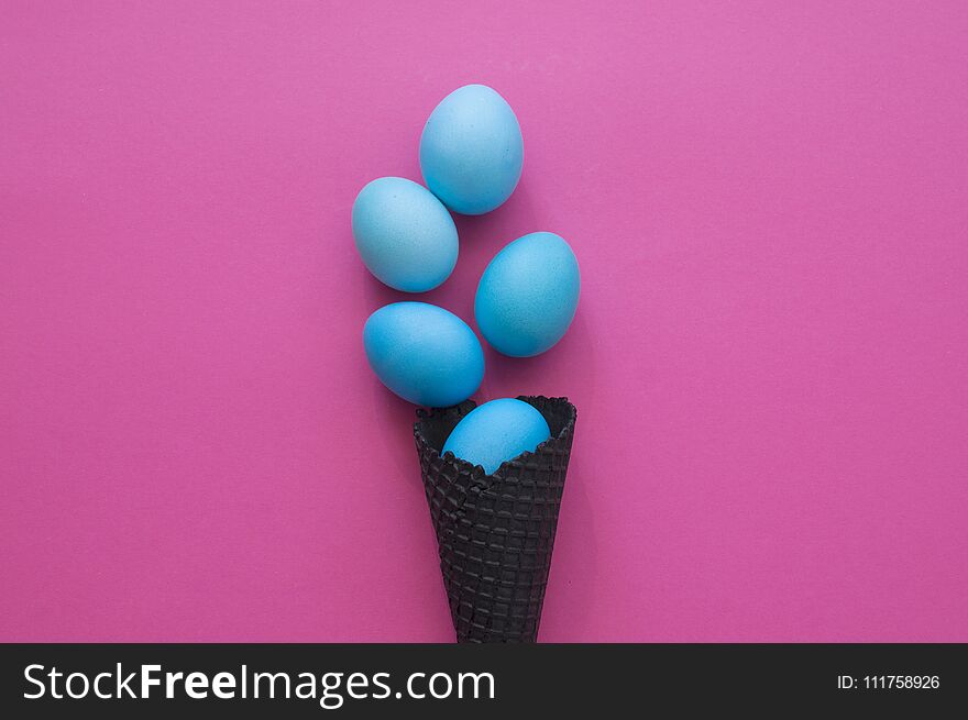 Easter. Blue eggs are poured from a waffle cone from ice cream. Black waffle cone. Ice cream egg. Pink background. Conceptual photo.