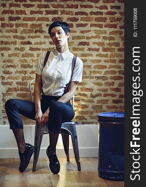 Beautiful young woman, model of fashion, with very short haircut, looking at camera sitting on chair, with a brick wall in the background. Beauty and hairstyles concept.