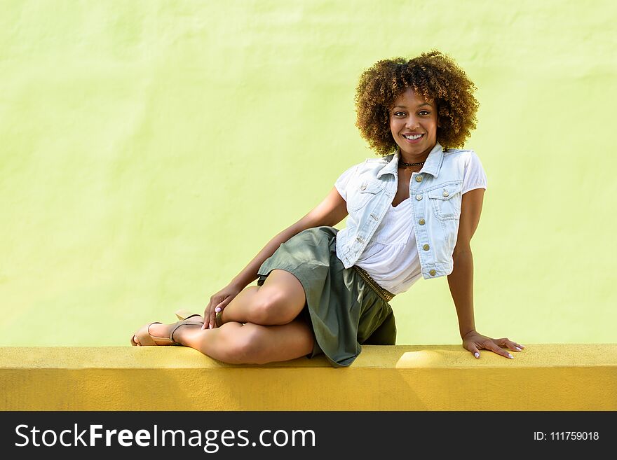 Young black woman, afro hairstyle, sitting on a wall smiling. Girl, model of fashion, wearing casual clothes in urban background. Female with skirt, denim vest and high heels. Young black woman, afro hairstyle, sitting on a wall smiling. Girl, model of fashion, wearing casual clothes in urban background. Female with skirt, denim vest and high heels.