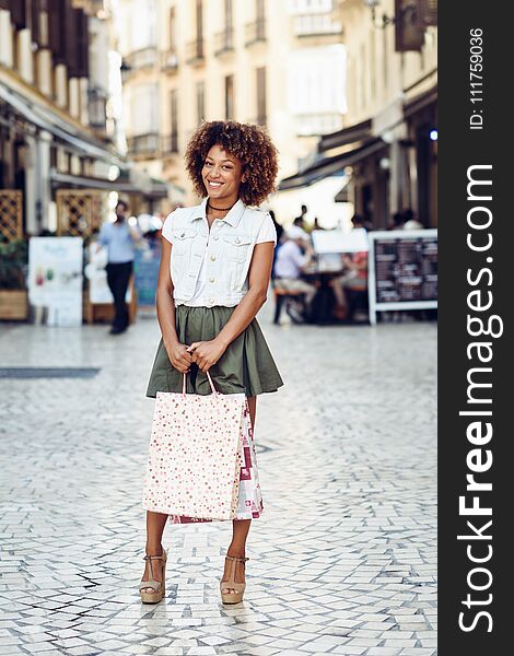 Young black woman, afro hairstyle, with shopping bags in the street. Girl wearing casual clothes in urban background. Female with skirt, denim vest and high heels.