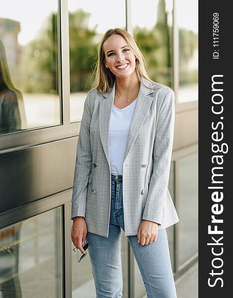 Beautiful young caucasian woman smiling in urban background. Blond girl wearing casual clothes in the street. Female with elegant jacket and blue jeans. Beautiful young caucasian woman smiling in urban background. Blond girl wearing casual clothes in the street. Female with elegant jacket and blue jeans
