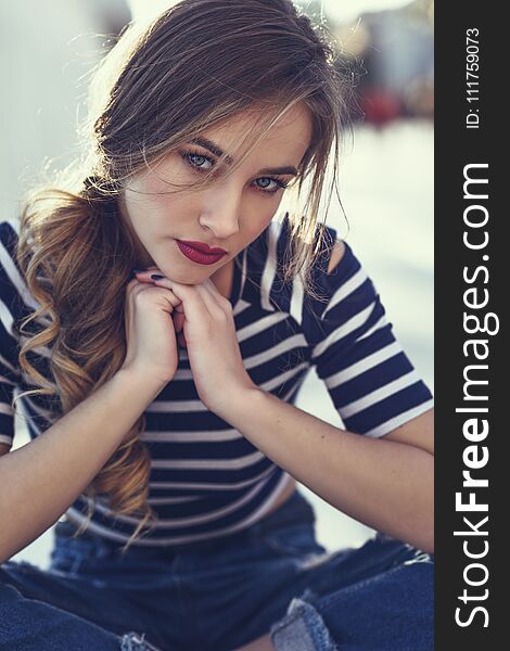 Blonde woman, model of fashion, sitting on a bench in urban background. Beautiful young girl wearing striped t-shirt and blue jeans in the street. Pretty russian female with pigtail. Blonde woman, model of fashion, sitting on a bench in urban background. Beautiful young girl wearing striped t-shirt and blue jeans in the street. Pretty russian female with pigtail.