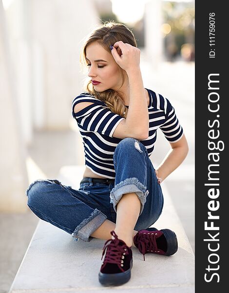 Blonde woman, model of fashion, sitting on a bench in urban background with eyes closed. Thoughtful young girl wearing striped t-shirt and blue jeans in the street. Pretty russian female with pigtail. Blonde woman, model of fashion, sitting on a bench in urban background with eyes closed. Thoughtful young girl wearing striped t-shirt and blue jeans in the street. Pretty russian female with pigtail.