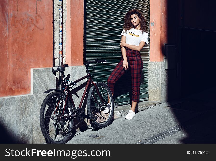 Beautiful young arabic woman with black curly hairstyle. Arab girl in casual clothes in the street near a bicycle. Girl power concept. Beautiful young arabic woman with black curly hairstyle. Arab girl in casual clothes in the street near a bicycle. Girl power concept.