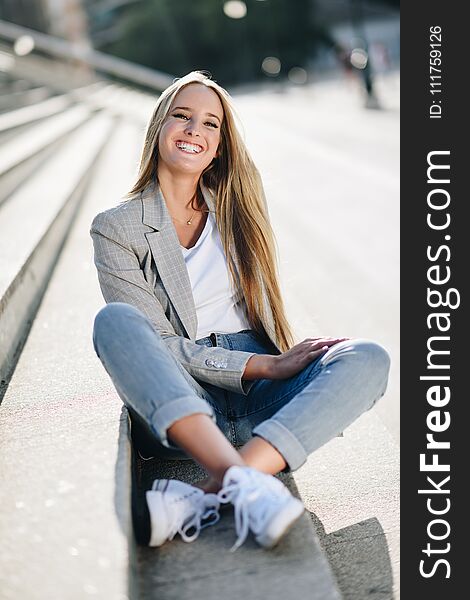 Beautiful young caucasian woman smiling in urban background. Blond girl wearing casual clothes in the street. Female with elegant jacket and blue jeans sitting on stairs. Beautiful young caucasian woman smiling in urban background. Blond girl wearing casual clothes in the street. Female with elegant jacket and blue jeans sitting on stairs.