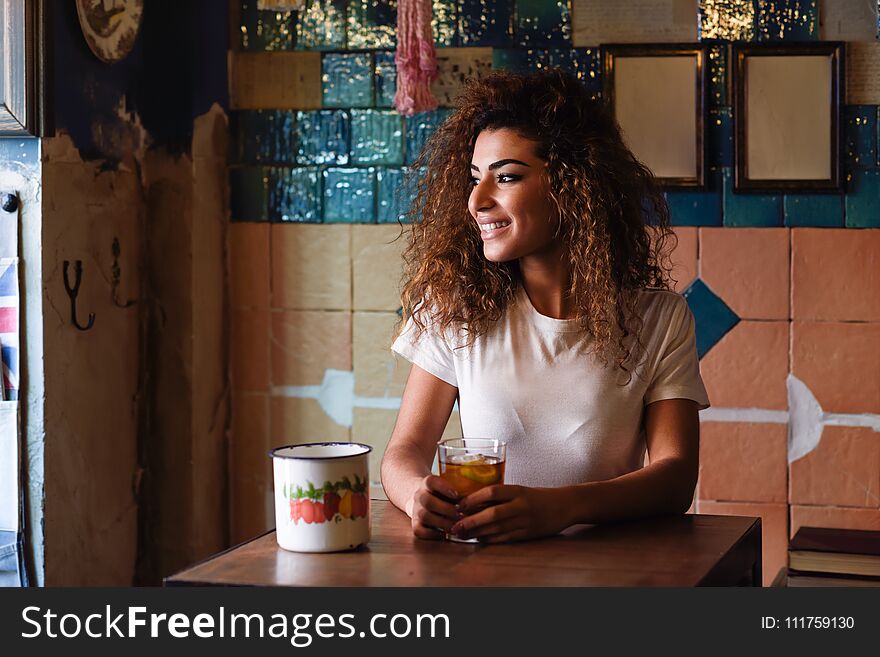 Young arabic smiling woman with black curly hairstyle sitting in a beautiful bar with vintage decoration. Arab girl in casual clothes drinking a soda. Young arabic smiling woman with black curly hairstyle sitting in a beautiful bar with vintage decoration. Arab girl in casual clothes drinking a soda.