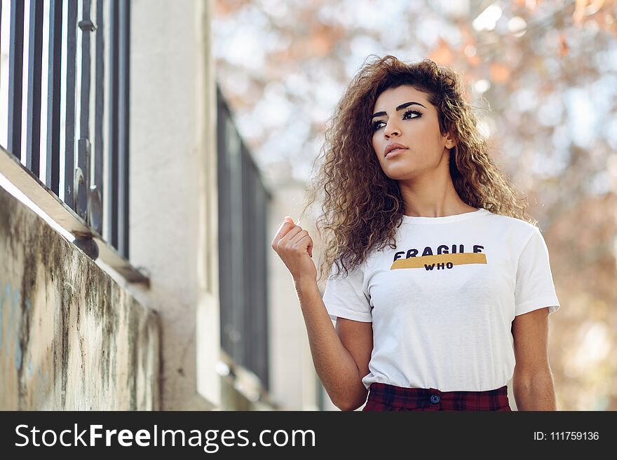 Beautiful young arabic woman with black curly hairstyle. Arab girl in casual clothes in the street. Happy female wearing white t-shirt and checked pants.