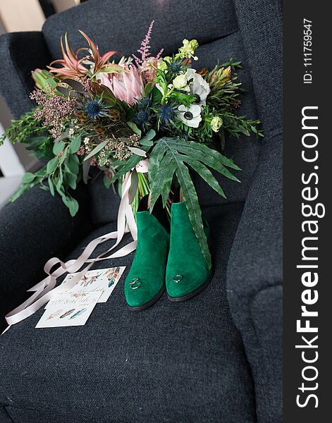 Rich green wedding bouquet with pink ribbons on the grey armchair. Green bridal shoes, and a wedding complimentary lying near the bridal lush bouquet. Details