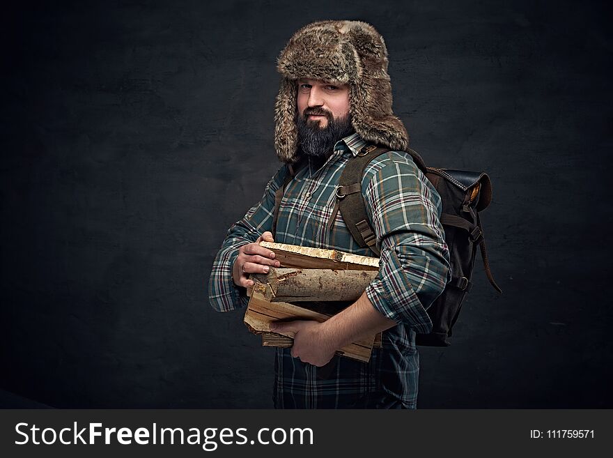 Bearded middle age male in a fur hat holds backpack and firewoods over dark grey background. Bearded middle age male in a fur hat holds backpack and firewoods over dark grey background.